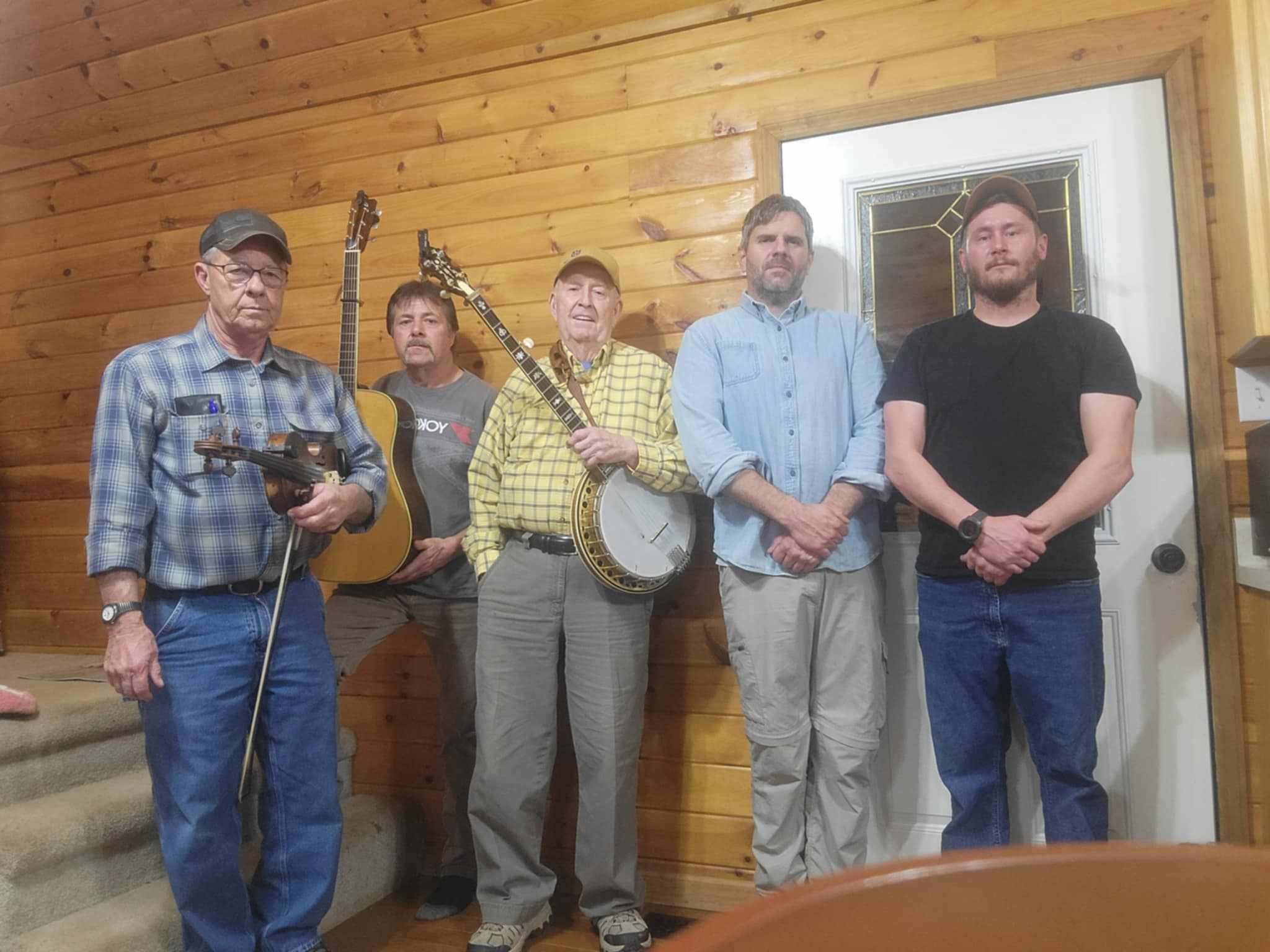 Five individuals stand together indoors, holding various musical instruments