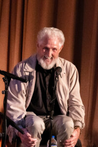 Man, Sonny Reighard, with white hair and beard smiling seated in front of a microphone.