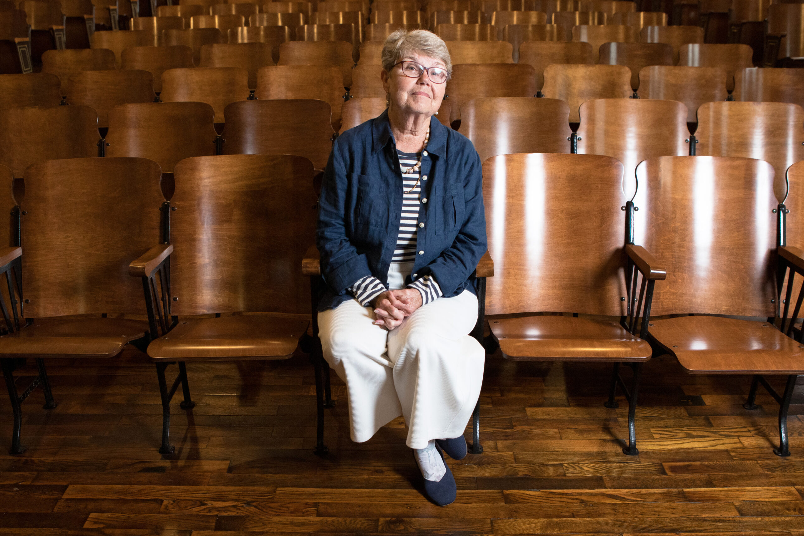 A portrait of Lynn Shields sitting in the front row of Stecoah Auditorium. Wide legged white pants, navy flats, striped shirt and blue linen jacket. Head tilted lights shining in her glasses.