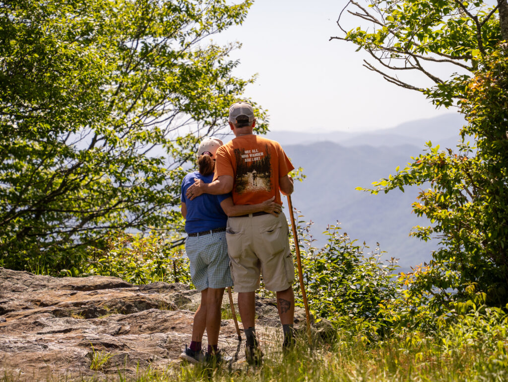 Two hikers standing on a rocky overlook, embracing while looking out at layered mountain ridges and lush green forest on top of Hooper Bald – one of the best hikes in Graham County, North Carolina.