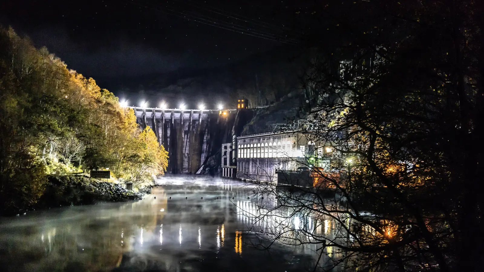 Cheoah Dam floodlit at night, its powerhouse lit from within along a row of tall arched windows, with mist rising off the tailwater below.