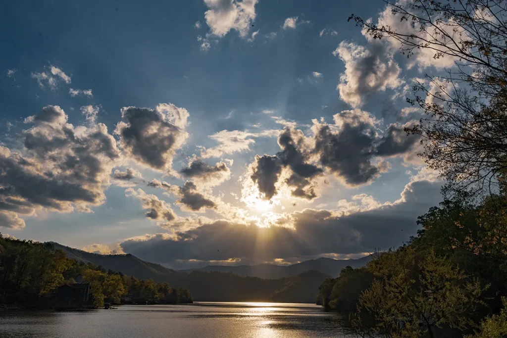 Sun breaking through scattered clouds over Lake Santeetlah at sunrise, casting rays across the water with forested ridges rising on either shore.