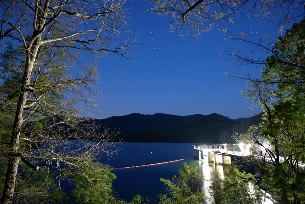 View of Santeetlah Dam from the overlook lit at night under a star-filled sky, with a line of red buoys crossing the dark water and forested ridges on the far shore.